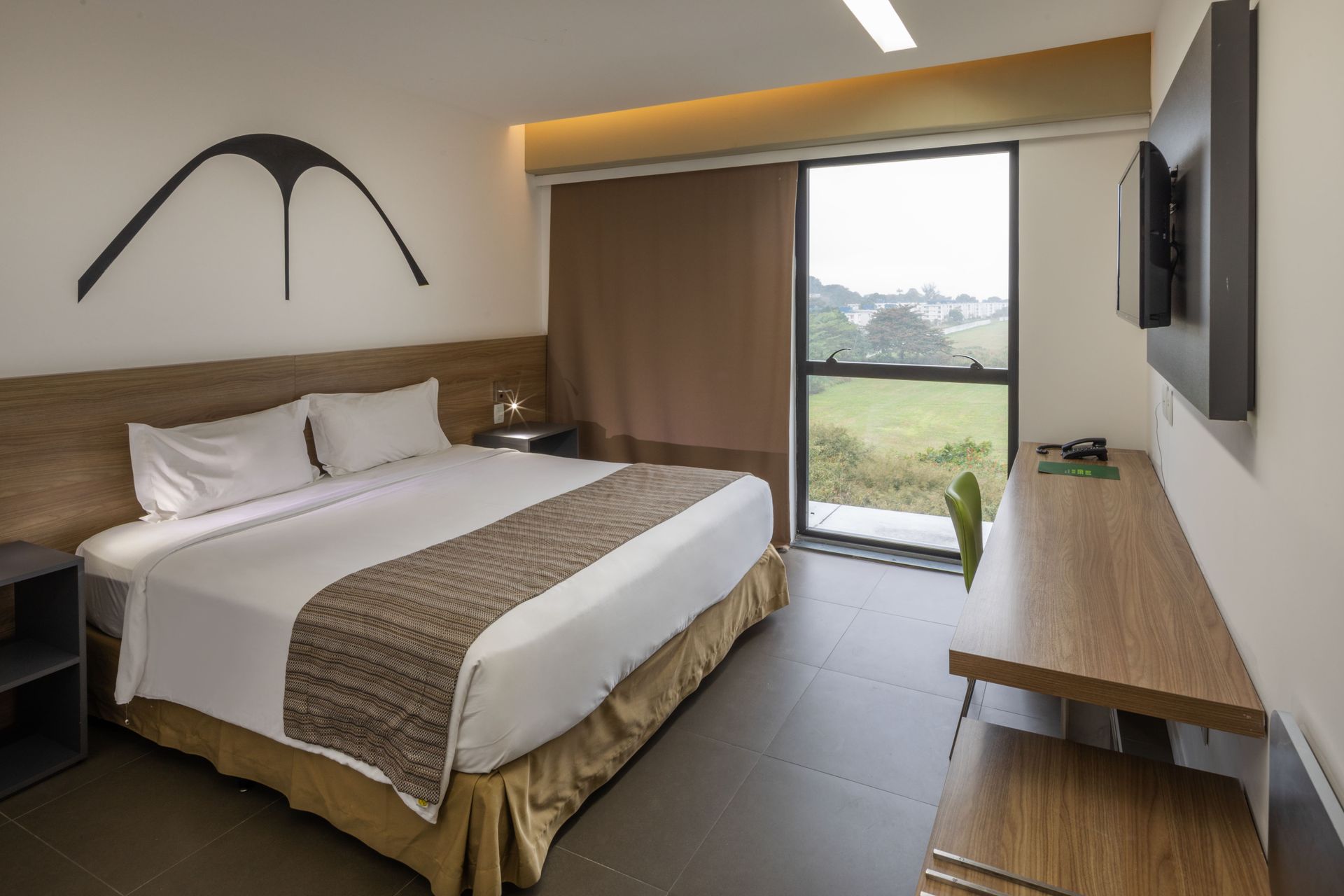 Quarto de hotel moderno com cama, escrivaninha, TV de parede e vista panorâmica para a área verde externa.