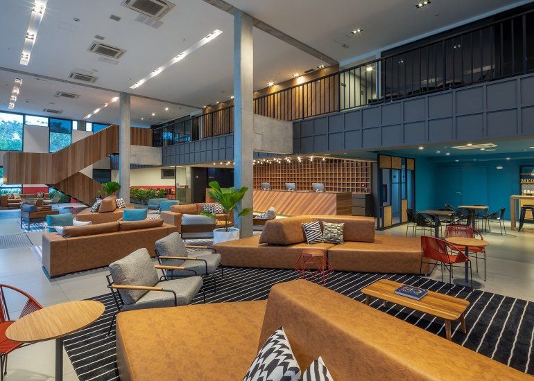 Sala de estar moderna com sofás bege, tapetes estampados, cadeiras azuis e um átrio com pé-direito alto.