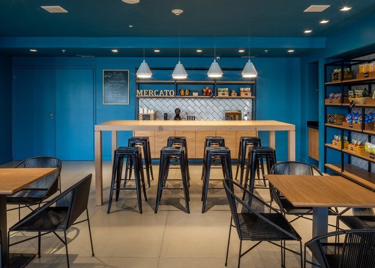 Interior de café moderno em tons de azul, com mesas de madeira, bancos pretos e luminárias pendentes no balcão de atendimento.