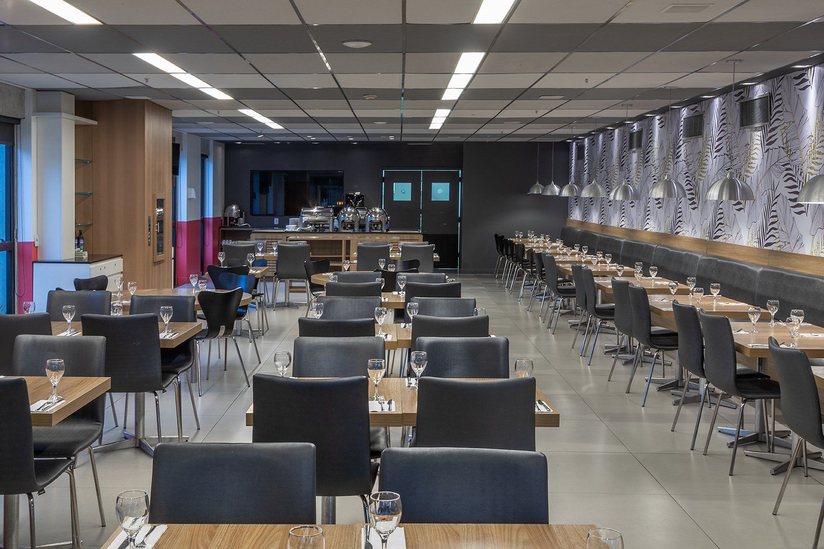 Restaurante vazio com fileiras de mesas e cadeiras sob luzes de teto, visto de frente.