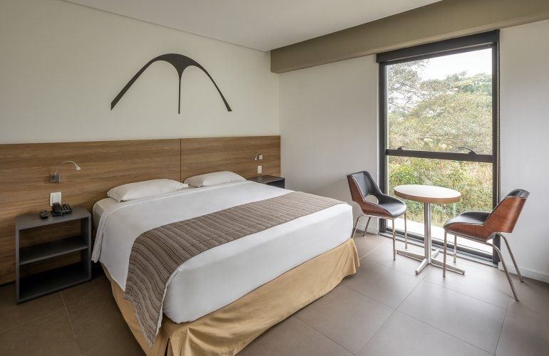 Quarto de hotel moderno com duas camas de solteiro, cabeceira de madeira e uma pequena mesa junto a uma grande janela.