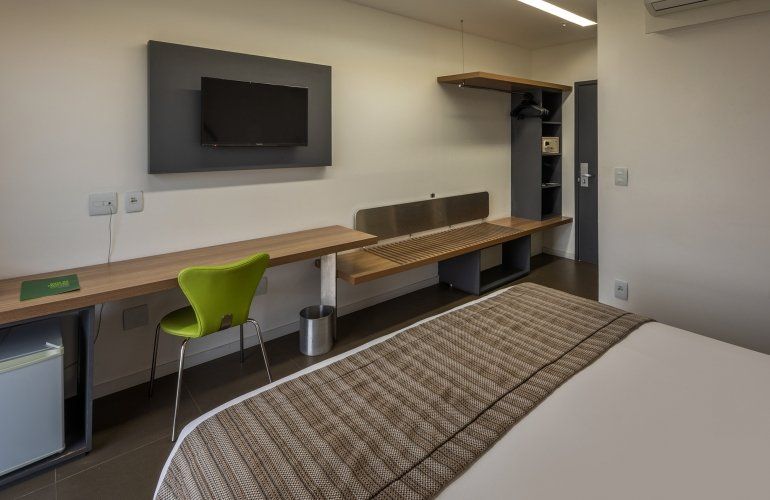 Quarto de hotel moderno com duas camas, TV de parede, escrivaninha, cadeira verde e banco junto à janela.
