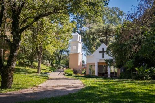 Pequena igreja branca com campanário ao lado de um caminho arborizado em um parque ensolarado.
