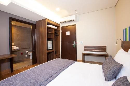Quarto de hotel compacto com cama, escrivaninha, guarda-roupa e porta do banheiro em tons de madeira aconchegantes.