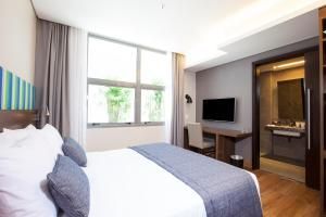 Quarto de hotel luminoso com cama, janelas amplas, TV, escrivaninha e banheiro integrado.