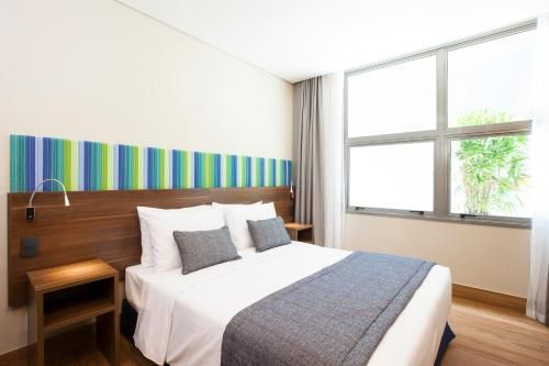 Quarto moderno com cama, cabeceira listrada, móveis de madeira e janela grande.