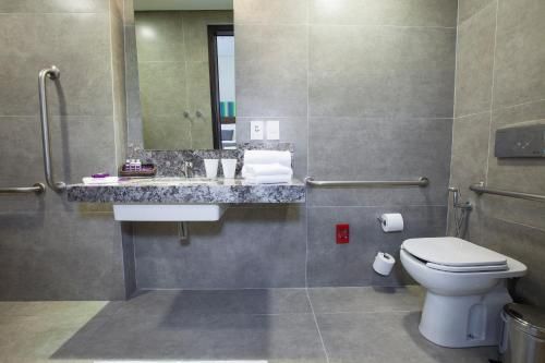 Banheiro acessível com azulejos cinza, pia, espelho, barras de apoio e vaso sanitário ao lado do box do chuveiro.