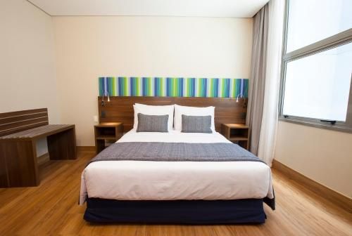 Quarto moderno com cama de casal, cabeceira listrada, escrivaninha e janela grande com cortinas.