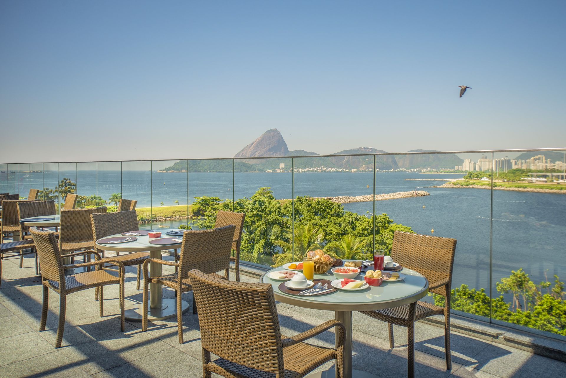 Mesas ao ar livre em um café à beira-mar com vista para uma baía azul e uma montanha ao longe sob um céu claro.