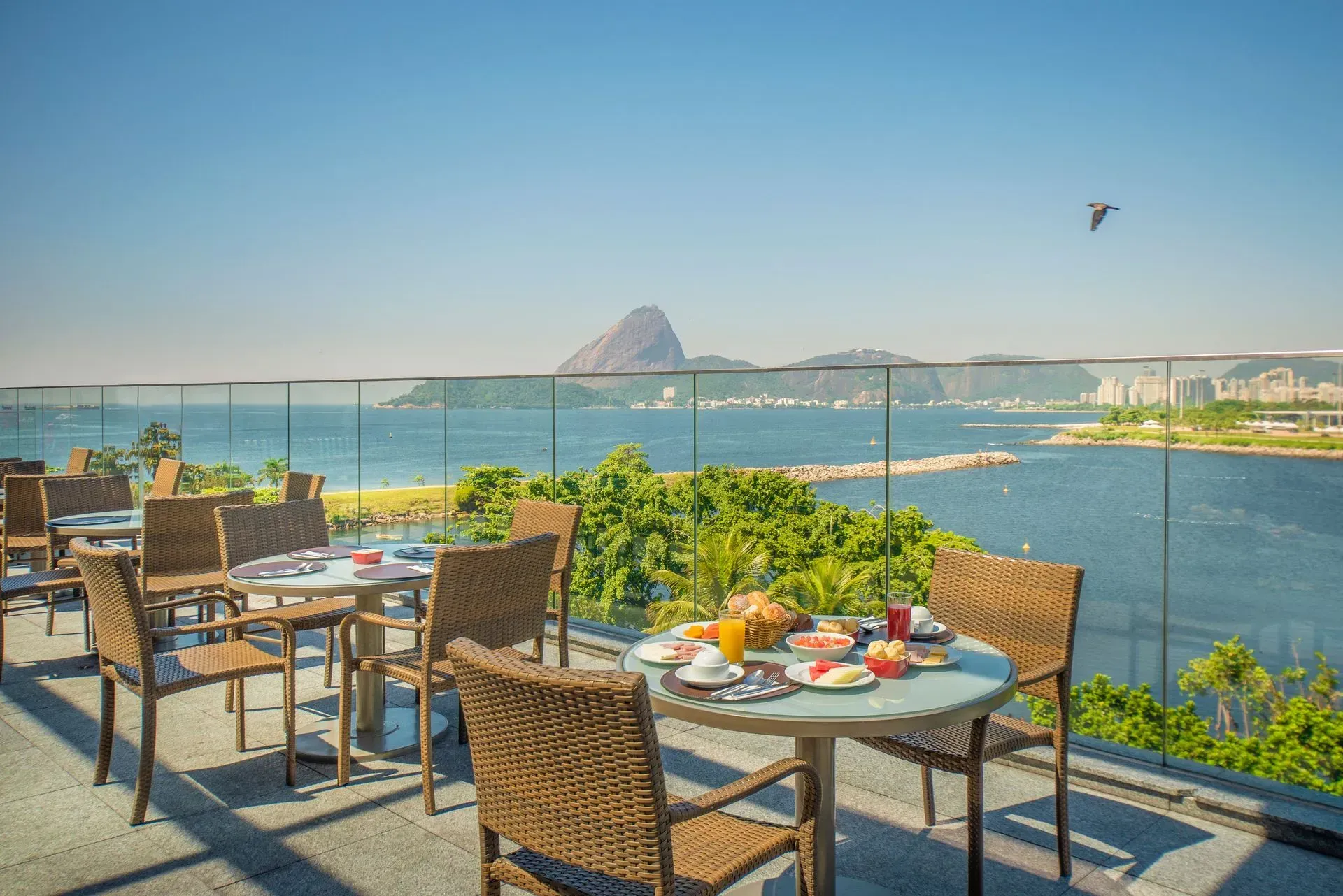 Terraço de restaurante à beira-mar com mesas com vista para uma baía azul e uma montanha ao longe sob um céu claro.