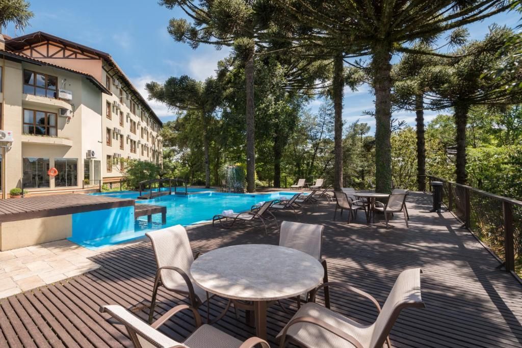 Pátio da piscina do resort com mesas, cadeiras e palmeiras ao lado de um hotel e piscina azul.
