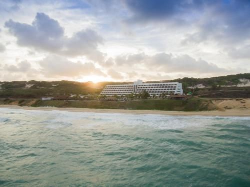 Pôr do sol sobre um resort costeiro com ondas em primeiro plano e céu nublado acima.