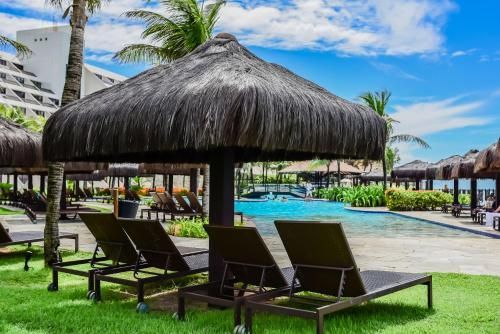 Espreguiçadeiras à beira da piscina do resort, sob guarda-sóis de palha, ao lado de uma piscina azul e palmeiras.