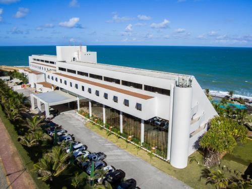 Hotel moderno e branco à beira-mar, com palmeiras e carros estacionados sob um céu azul.