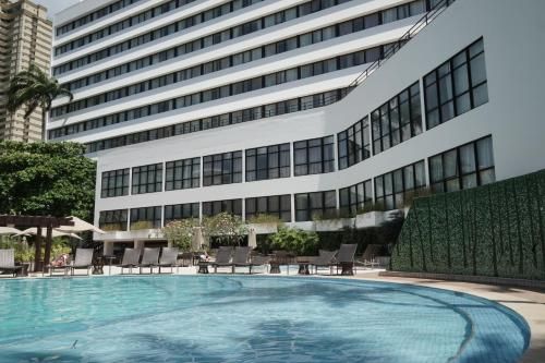 Piscina curva de hotel ao lado de um prédio branco moderno com espreguiçadeiras e vegetação.