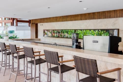 Bar moderno no lobby do hotel com banquetas de vime, balcão longo de mármore e parede verde ao fundo.
