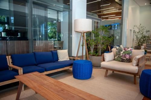 Sala de estar moderna com sofá azul, poltrona bege, mesa de centro de madeira e plantas em vasos em um saguão iluminado com paredes de vidro.