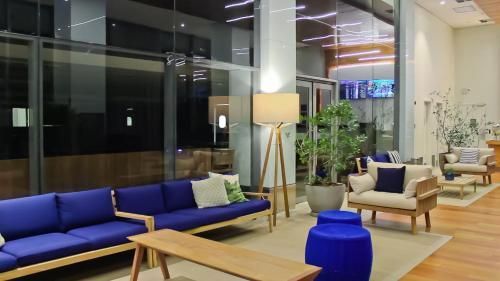 Hall de entrada moderno com sofás azuis, poltronas, um abajur de chão, plantas e uma mesa de madeira junto a grandes janelas de vidro.