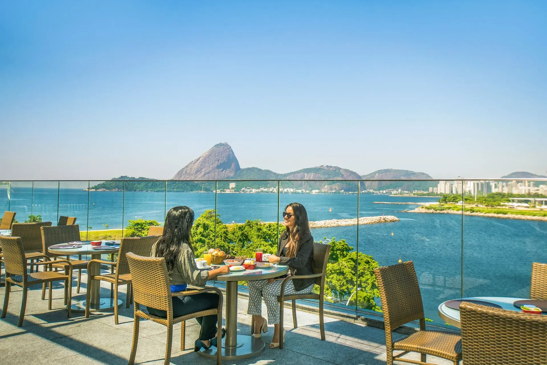 Pessoas jantando à beira da água com vista para a montanha e céu azul.