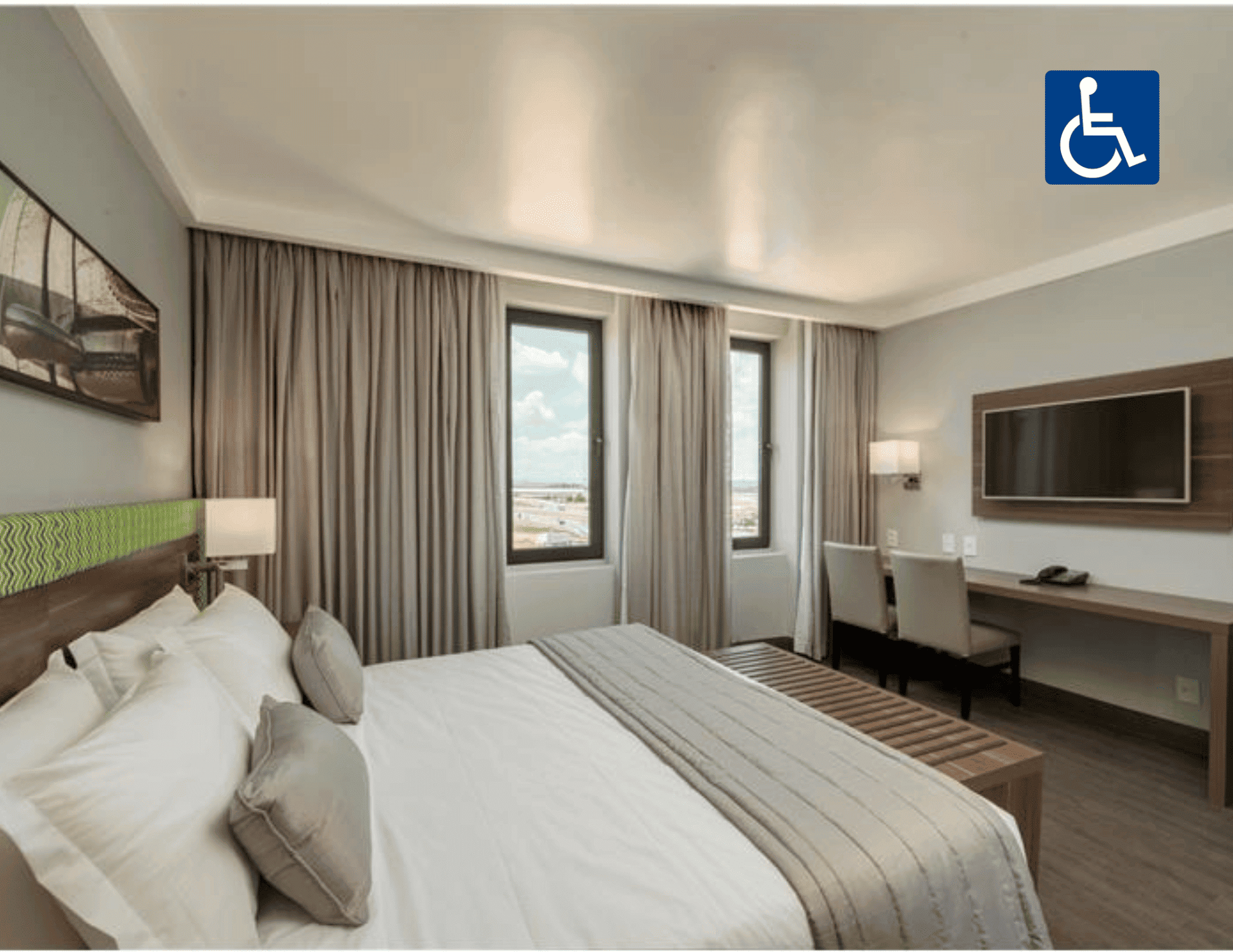 Quarto de hotel acessível com cama, escrivaninha, TV e janela ampla com vista para a cidade.