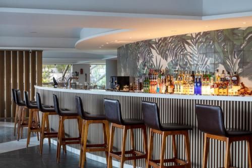 Interior de bar moderno com banquetas altas, garrafas iluminadas e um mural de parede com estampa de folhas.