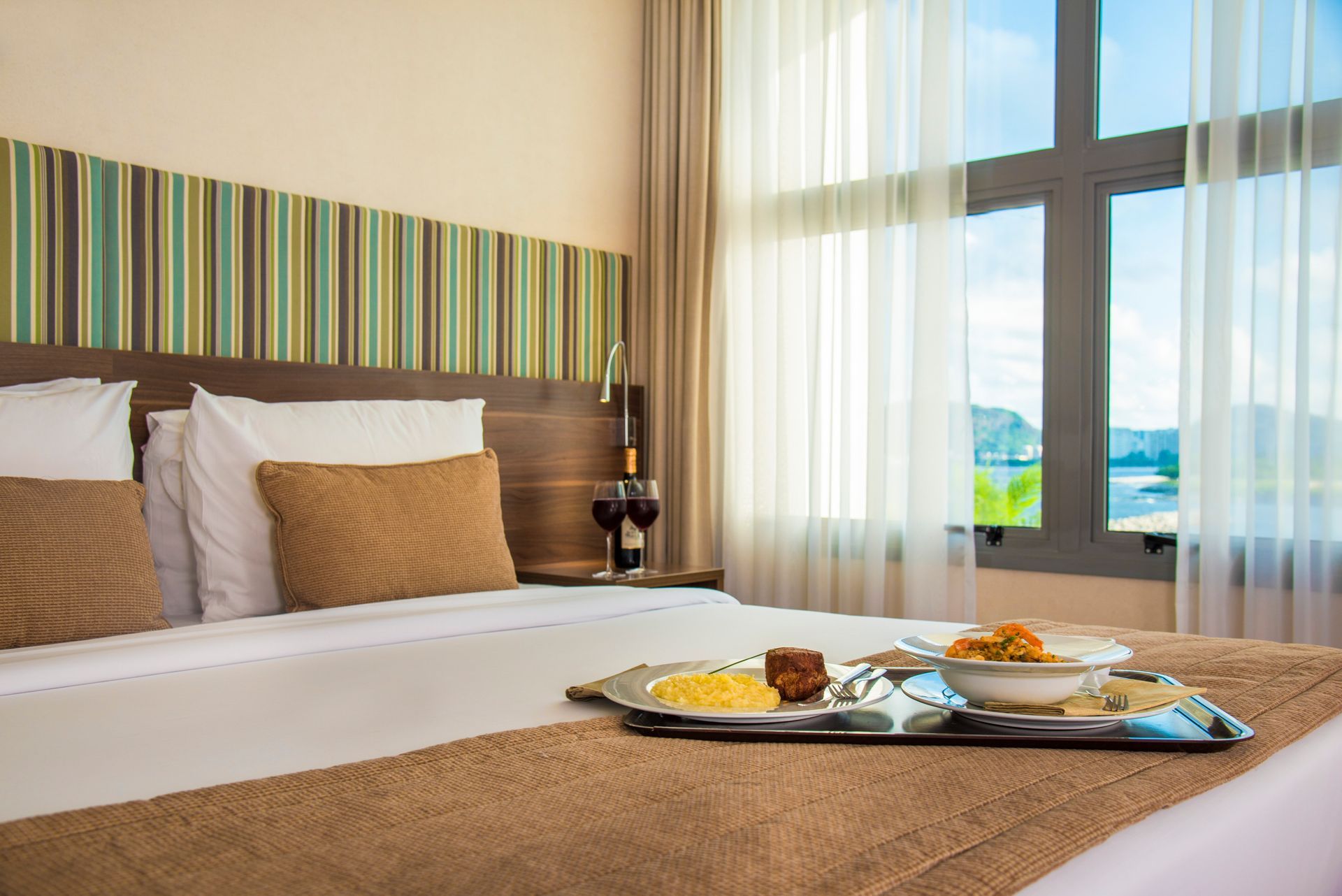 Quarto de hotel com cama bem arrumada, bandeja de café da manhã e vista para o mar pela janela.