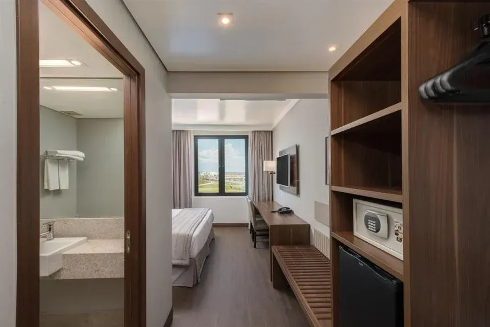 Quarto de hotel moderno com cama, escrivaninha, TV e armário aberto, visto da entrada do banheiro.