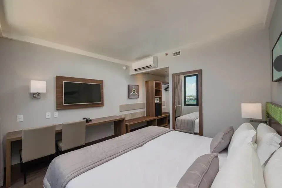Quarto de hotel moderno com cama, escrivaninha, TV de parede e porta aberta para o banheiro.