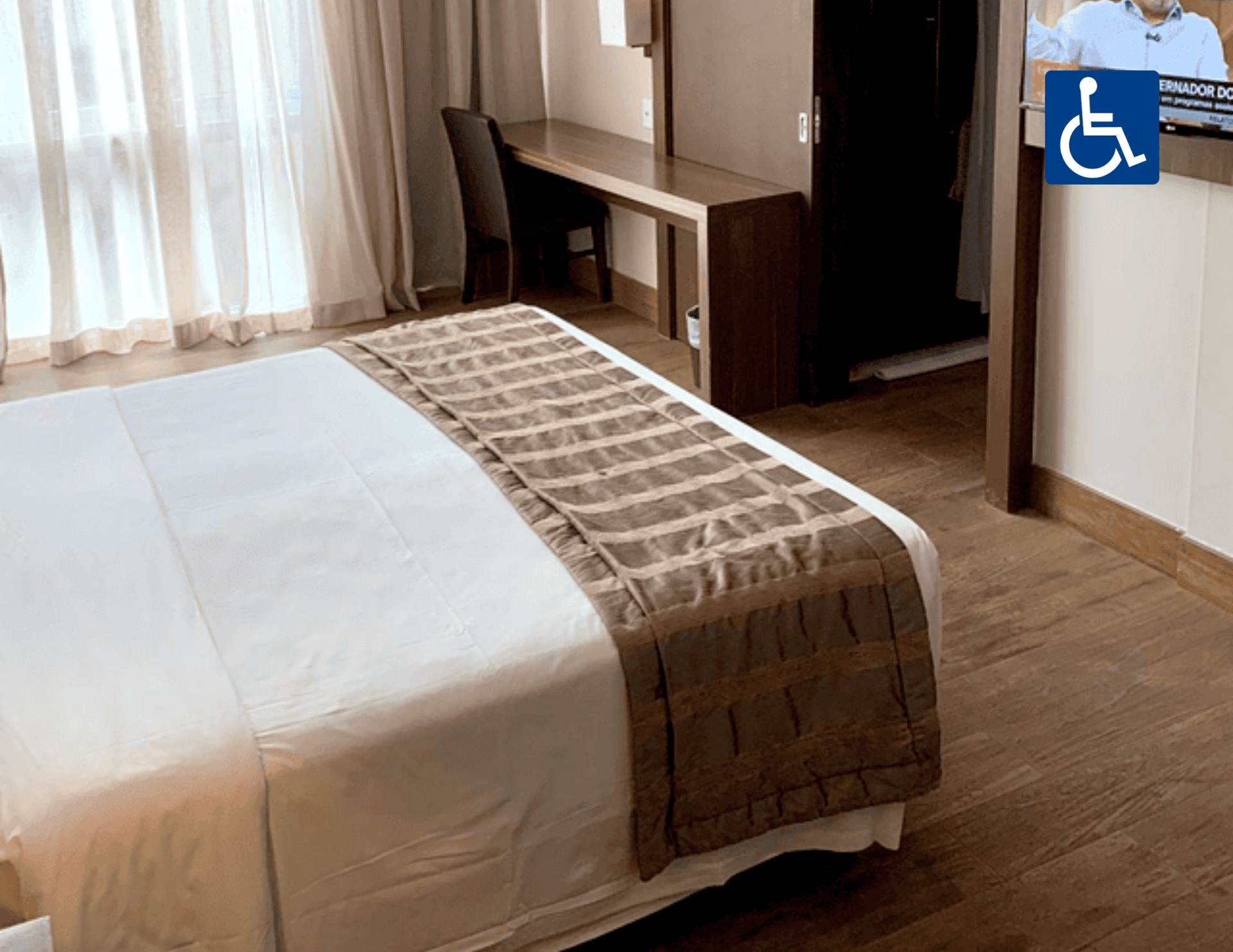 Quarto de hotel acessível com cama arrumada, escrivaninha junto à janela e placa de acessibilidade para cadeirantes na parede.