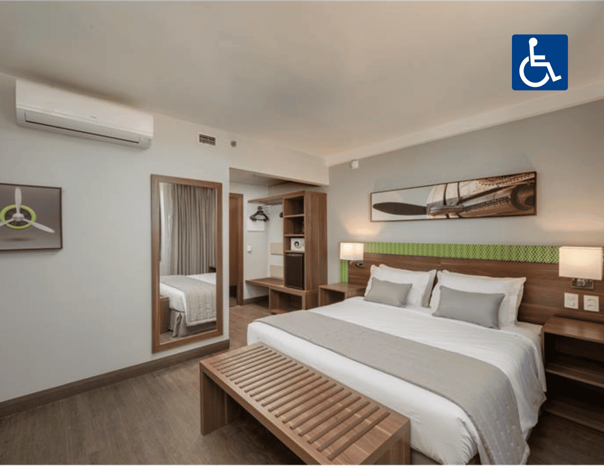 Quarto de hotel acessível com cama king-size, banco, espelho de parede, ar-condicionado e porta para o banheiro.
