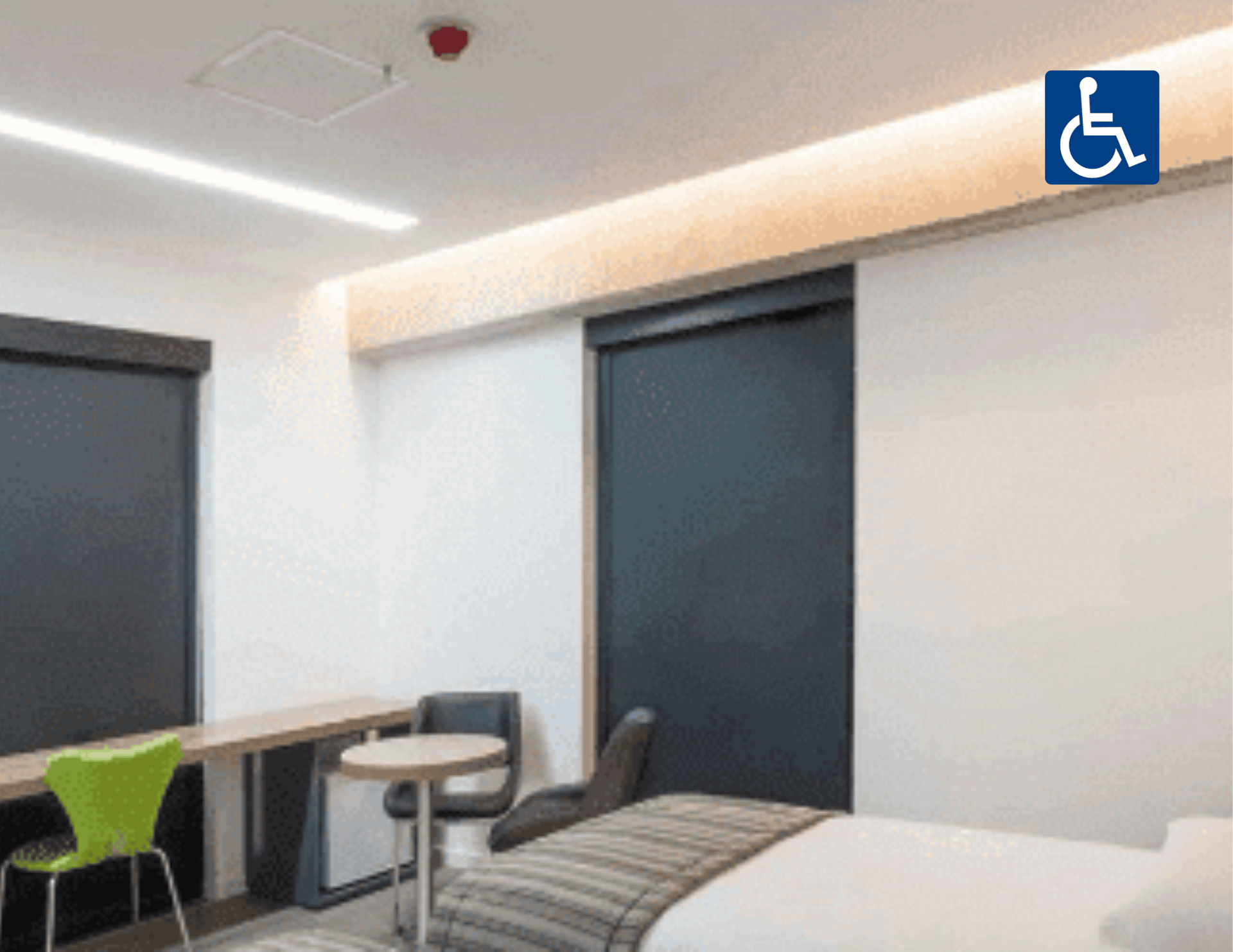 Quarto de hotel acessível com cama, escrivaninha, cadeira e placa azul de cadeira de rodas perto da porta.