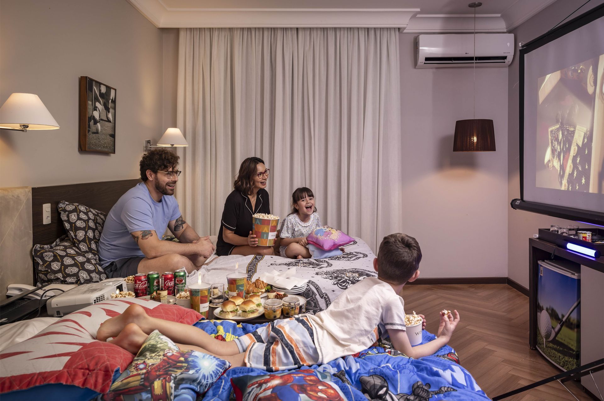 Família sentada em camas num quarto aconchegante, assistindo TV e comendo petiscos numa bandeja compartilhada.