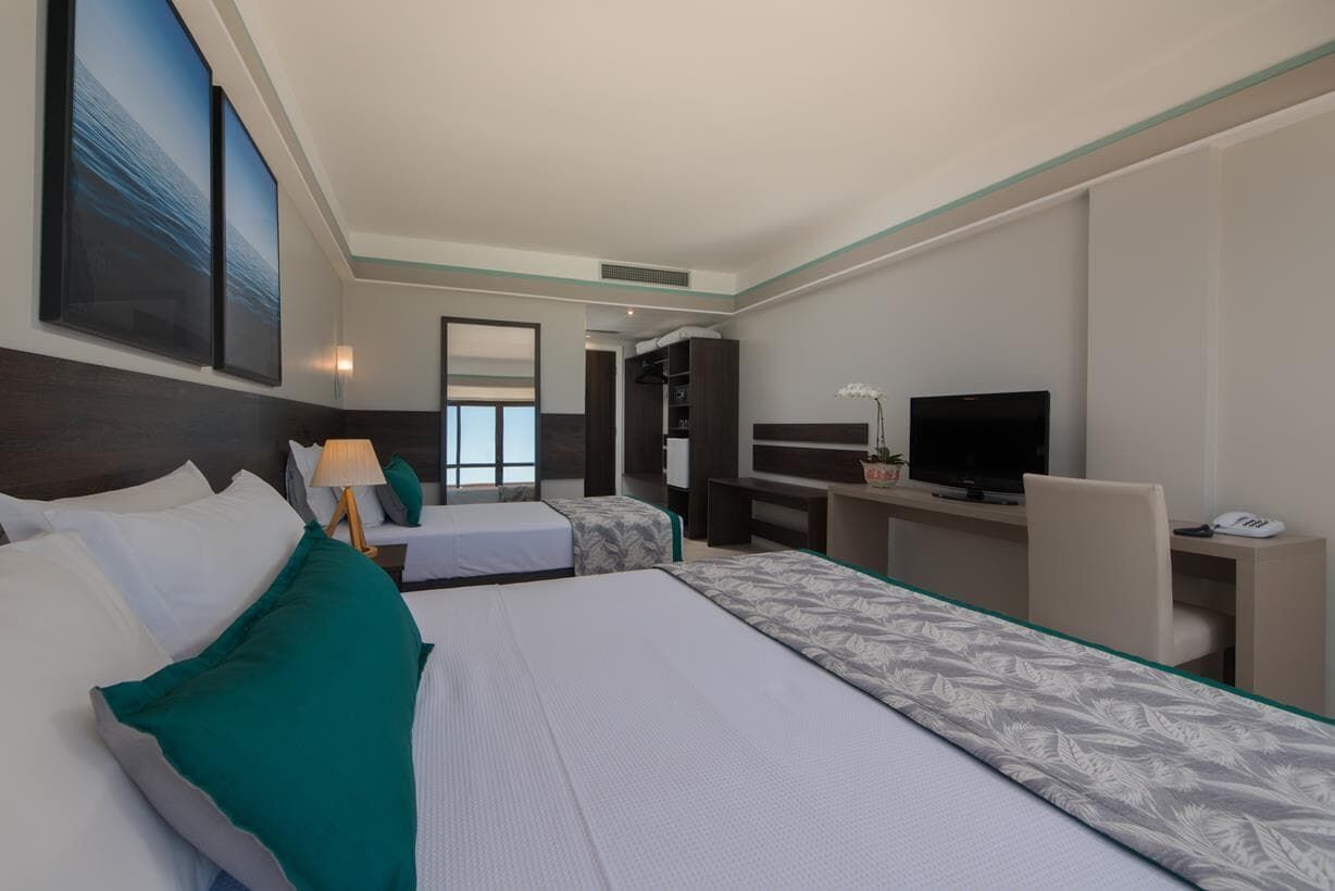 Quarto de hotel moderno com duas camas, TV, escrivaninha e janelas amplas.