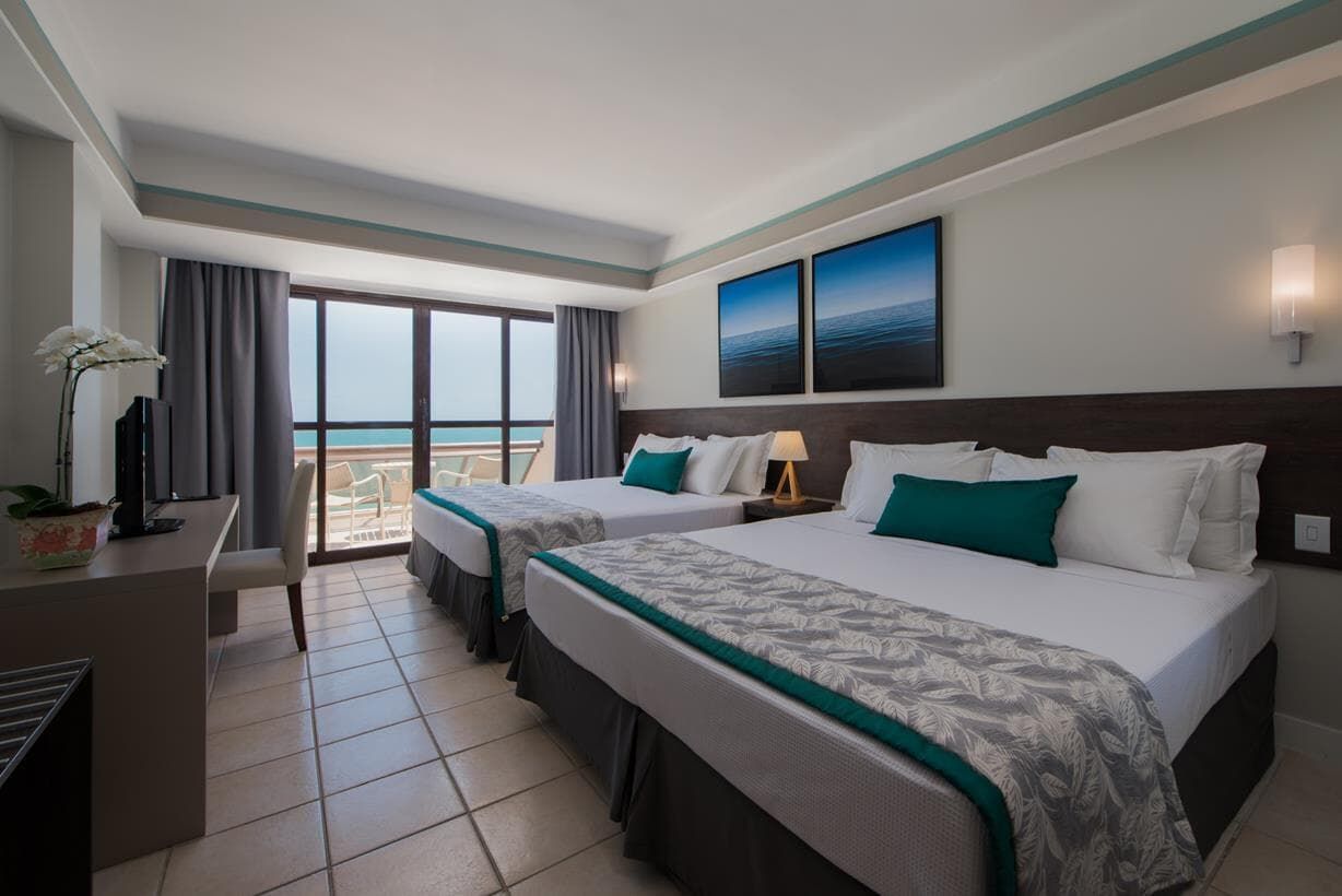 Quarto de hotel moderno com duas camas, varanda com vista para o mar e detalhes em azul-turquesa.