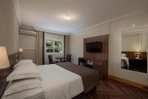 Quarto de hotel moderno com cama grande, TV de parede, janela e espelho sobre piso de madeira.
