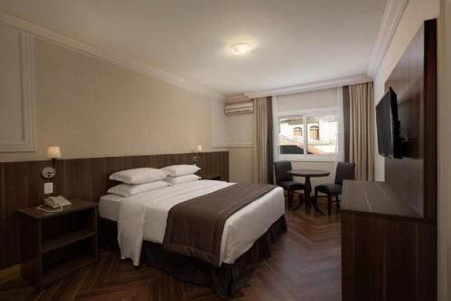 Quarto de hotel moderno com cama bem arrumada, escrivaninha, TV de parede e área de estar junto à janela.