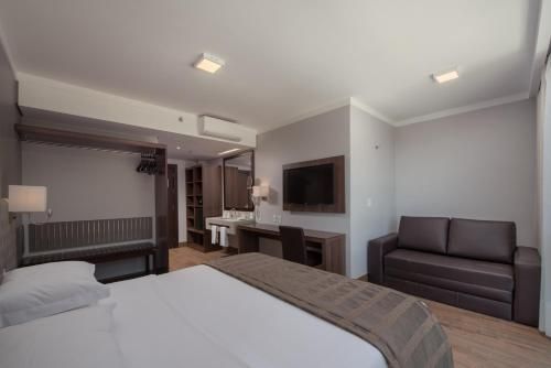 Quarto de hotel moderno com duas camas, sofá cinza, escrivaninha e TV em um interior luminoso.