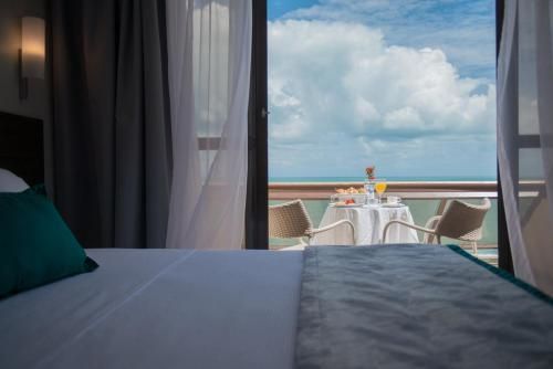 Vista do quarto de hotel para uma mesa na varanda com vista para o oceano, com cortinas emoldurando a paisagem e um céu azul.