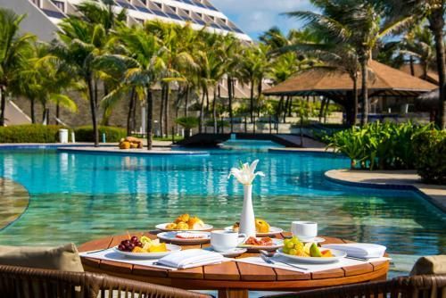 Bandeja de café da manhã à beira da piscina turquesa do resort, com palmeiras e espreguiçadeiras.