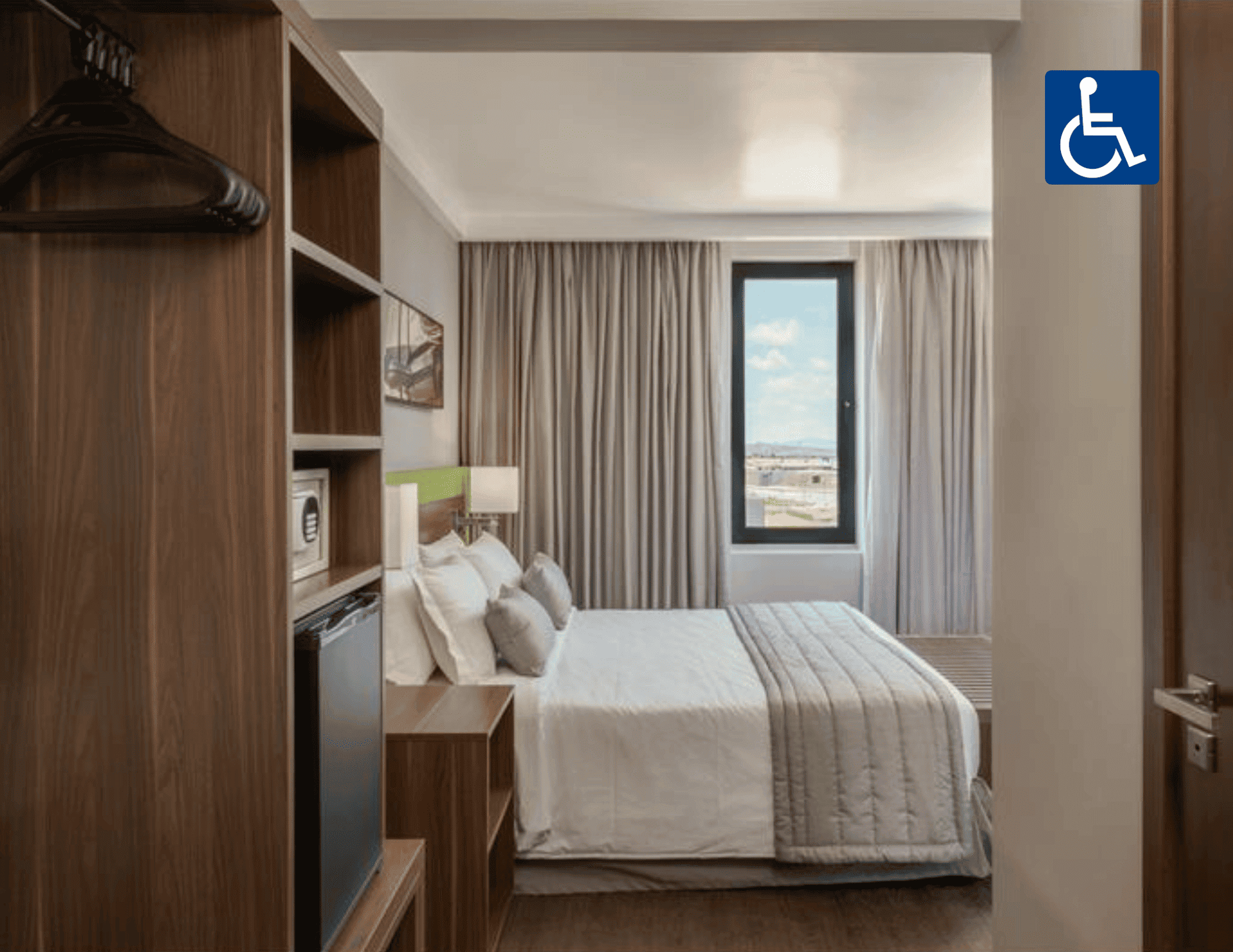 Quarto de hotel acessível com cama arrumada, cortinas, janela e nicho para guarda-roupa.