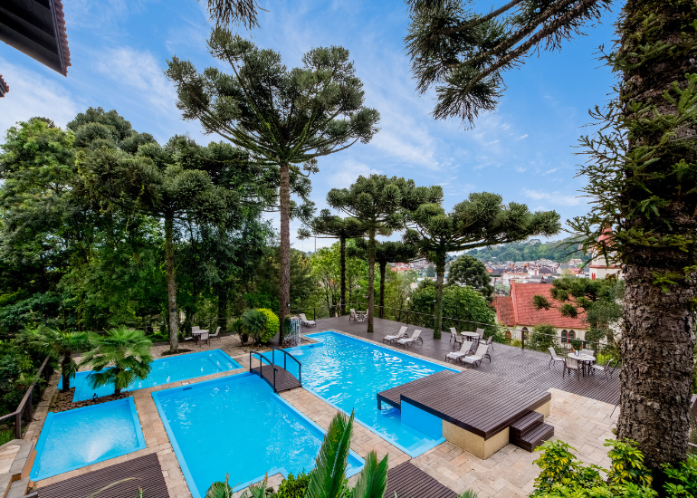 Área da piscina do resort com piscinas de água turquesa, espreguiçadeiras e pinheiros com vista para uma cidade costeira.