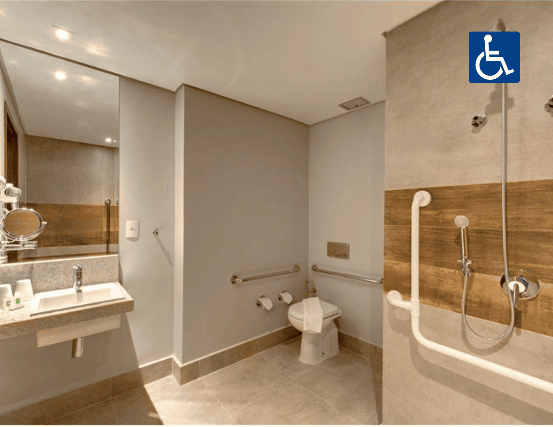 Banheiro acessível no hotel com chuveiro de acesso facilitado, vaso sanitário, pia e barras de apoio.