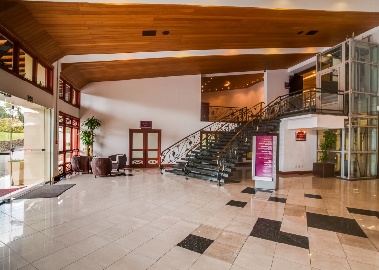 Amplo hall de entrada do hotel com piso de azulejo, teto de madeira, escadaria e elevador de vidro.