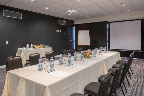 Sala de conferências com mesas compridas, garrafas de água, cadeiras pretas e tela de projeção.