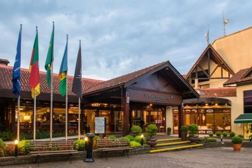 Entrada moderna de hotel ao entardecer, com bandeiras, luzes aconchegantes e um saguão com teto de telhas.