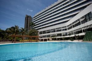 Grande hotel moderno com fachada curva ao lado de uma piscina turquesa e palmeiras.