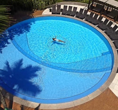Piscina circular azul com uma pessoa flutuando no centro, rodeada por espreguiçadeiras.