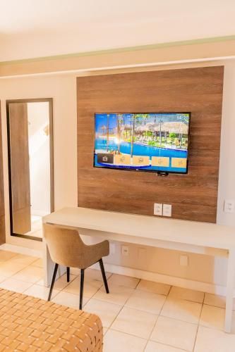 Quarto pequeno com uma TV fixada na parede acima de uma mesa e cadeira, ao lado de um espelho de corpo inteiro e piso de azulejo.