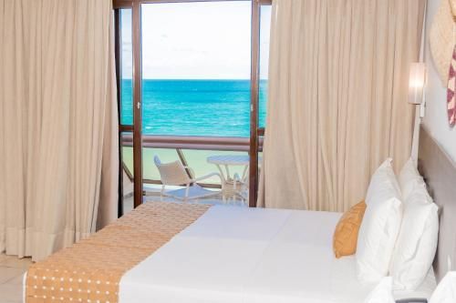 Quarto de hotel com portas de varanda abertas com vista para o oceano turquesa e duas espreguiçadeiras.