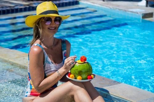Mulher de chapéu amarelo sentada à beira de uma piscina, segurando um prato de frutas e sorrindo.